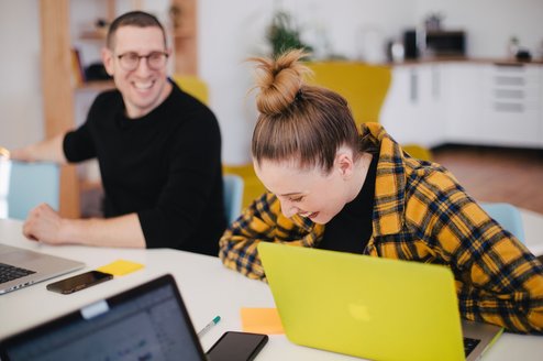two students laughing while sitting at a desk in front of laptops